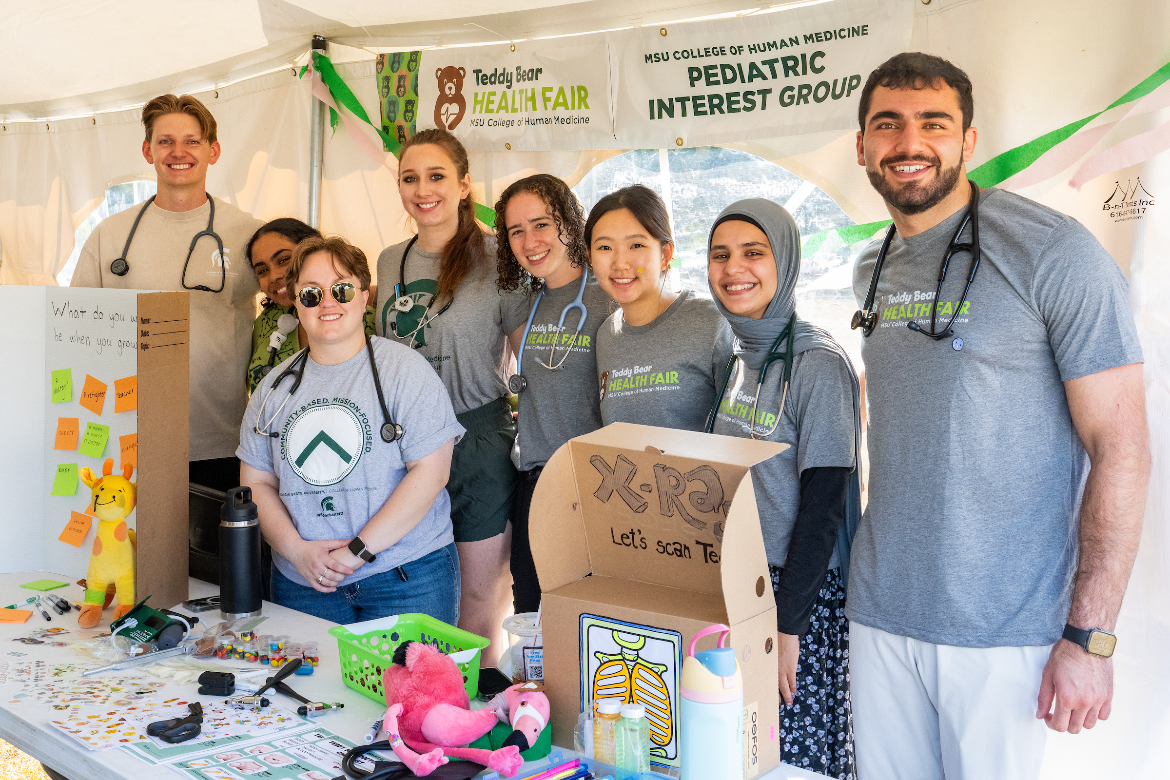 MSU College of Human Medicine students at the Teddy Bear Health Fair event in Grand Rapids. They are standing in front of a table with a kid friendly "x-ray" machine.