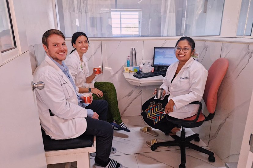 Three people in a small office, wearing white lab coats, smiling and holding mugs next to a computer.